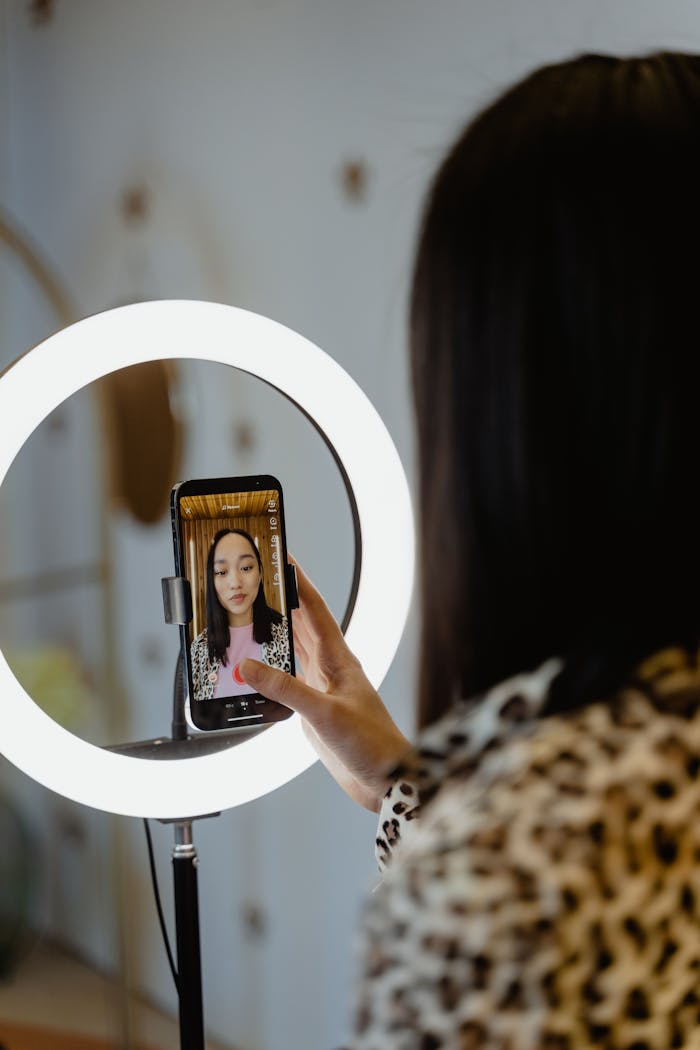Young woman recording video with smartphone and ring light indoors.